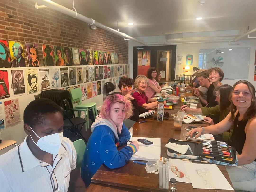 Drawing Club around a long table in a brick-walled studio hung with hand-made posters, everyone looking up from their sketchbooks.