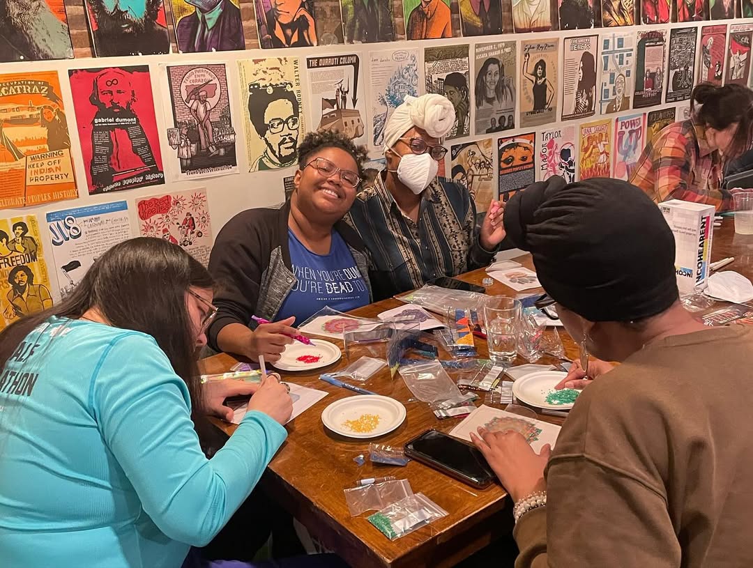 Drawing Club members beading and drawing at a wooden table, laughing, with a wall of political posters behind them.